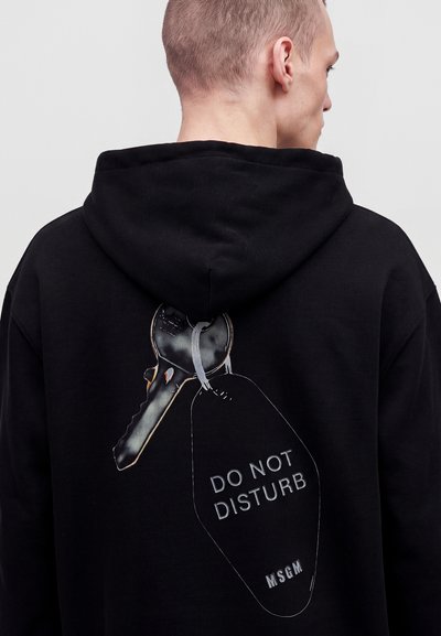 Homme vu de dos portant un sweat à capuche noir avec une grande clé et un graphisme d’étiquette "Do Not Disturb MSGM" dans le dos.