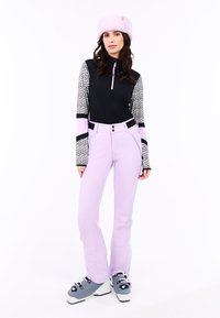 Juodas ir raštuotas ilgomis rankovėmis viršus derinamas su levandų spalvos slidinėjimo kelnėmis. Modelis vilki pilkas slidinėjimo batus ir šviesiai rožinę firminę kepurę, stovintis ant baltos fono.