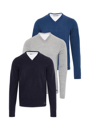 Homme portant un pull à col en V bleu, un sous-pull blanc, un jean foncé et des baskets blanches, debout devant un fond clair uni.