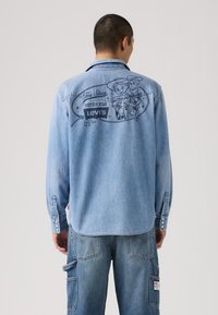 Lyseblå denimskjorte med lange ermer, med et grafisk trykk på ryggen av Toy Story-karakterer og Levi's merkevare.