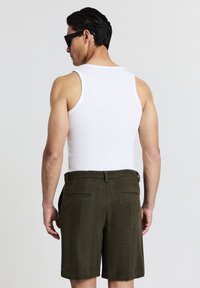 Homme portant des lunettes de soleil noires, un débardeur blanc sans manches et un short texturé vert foncé, se tenant de dos contre un fond uni.