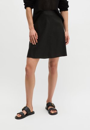 Person trägt einen schwarzen knielangen Rock, einen schwarzen Strickpullover und schwarze Sandalen mit goldenen Schnallen, steht vor einem schlichten weißen Hintergrund.