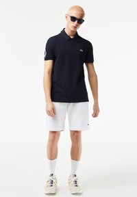 Marinblå pikétröja med korta ärmar, med en liten logotyp. Kombinerad med vita shorts och ljusa sneakers med nät detaljer och vita strumpor.