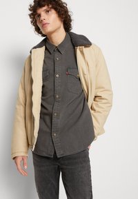 Chaqueta beige con cremallera y un cuello de piel sintética gris, sobre una camisa de botones gris oscuro, combinada con jeans gris oscuro; texturas suaves y diseño casual.