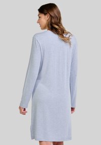 Mujer con cabello ondulado que viste un vestido largo de manga larga hasta la rodilla de color gris claro, de pie contra un fondo simple, mirando hacia atrás.