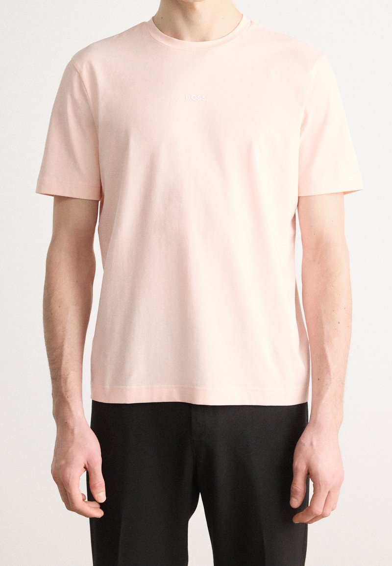 Homme portant un t-shirt à manches courtes rose clair avec un petit logo "BOSS" sur la poitrine et un pantalon noir sur un fond uni.