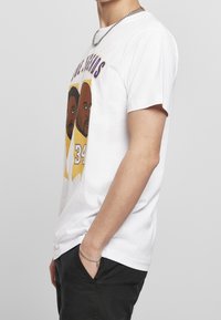 Man med vit T-shirt med illustrerade ansikten och nummer 34 i gult, svarta byxor, silverkedja som halsband och armband, händerna i fickorna.