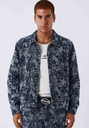 Young man wearing blue floral patterned denim jacket and pants, white shirt with black logo, silver chain necklace, and hoop earrings.
