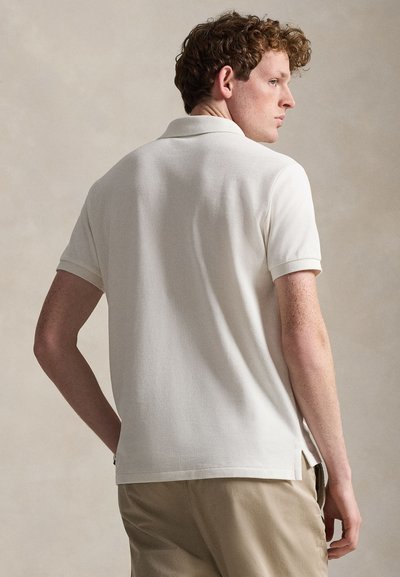 Νέος με κατσαρά μαλλιά που φορά λευκό κοντομάνικο πόλο πουκάμισο και μπεζ παντελόνι, γυρισμένος ελαφρώς στο πλάι μπροστά σε ουδέτερο φόντο.