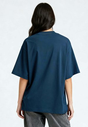 Woman with long dark hair wearing a loose dark blue short-sleeve shirt and gray jeans, facing away against a plain light background.