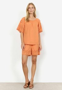 Top in cotone color pesco con maniche in broderie anglaise abbinato a pantaloni corti con orlo scallop. Sandali marroni con cinturini.