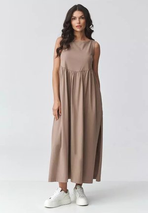 Kobieta z długimi ciemnymi kręconymi włosami, ubrana w beżową, bezrękawnikową suknię maxi oraz białe sneakersy na platformie, stoi na tle jasnego, jednolitego tła.