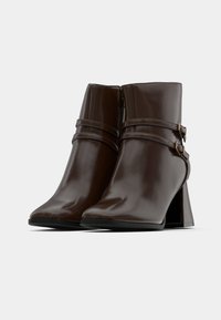 Botas de tobillo de cuero marrón con un acabado brillante, con tacones angulares, dos correas decorativas y hebillas de metal, punteras afiladas y cierre de cremallera.