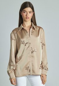 Camicia beige in satin con maniche lunghe, decorata con motivi floreali in paillettes, dotata di chiusura a bottoni sul davanti e colletto arrotondato.