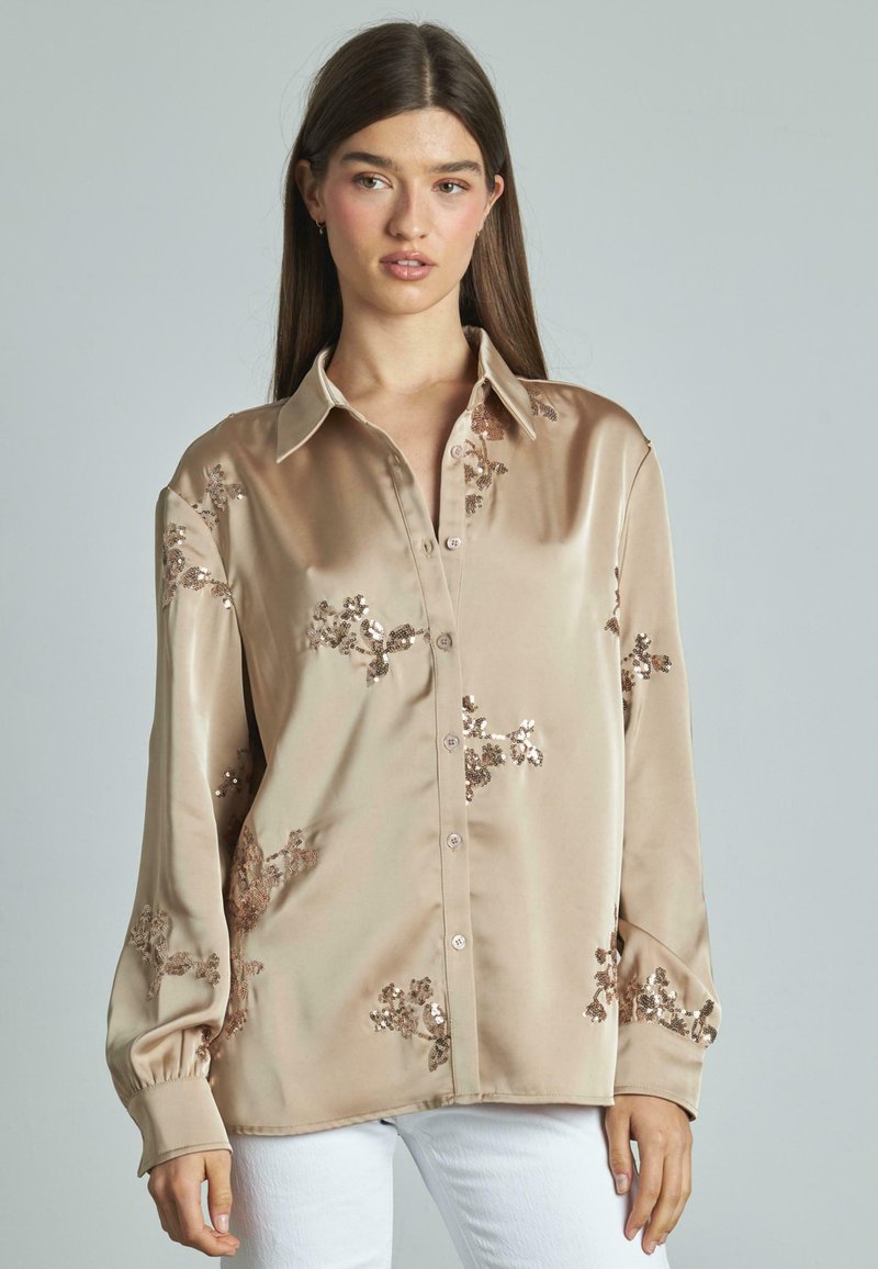 Camicia beige in satin con maniche lunghe, decorata con motivi floreali in paillettes, dotata di chiusura a bottoni sul davanti e colletto arrotondato.