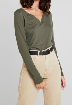 Femme portant un haut à manches longues vert olive rentré dans un pantalon beige taille haute avec une ceinture noire et une boucle dorée.
