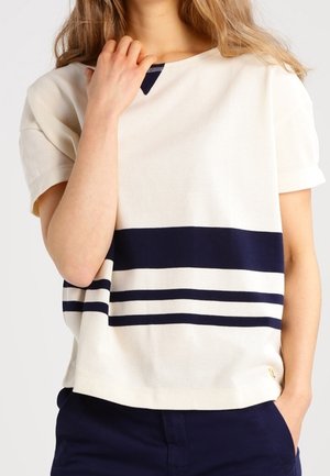 Jeune personne portant un haut crème à manches courtes avec trois bandes horizontales bleu marine sur la poitrine et un pantalon bleu marine.