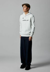 Jeune homme debout, portant un sweat à capuche gris clair avec le texte "athletic club", un jean large noir et des chaussures noires, contre un fond gris uni.