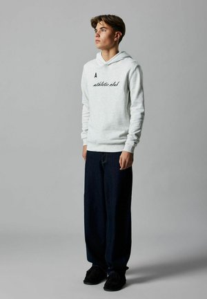 Jonge man staat, draagt een lichtgrijze hoodie met de tekst "athletic club", donkere wijde jeans en zwarte schoenen, tegen een effen grijze achtergrond.