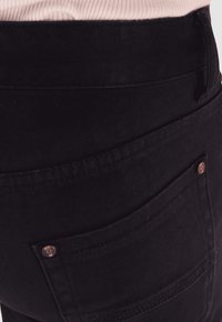 Vaqueros de mezclilla negra con una cinturilla alta, ajuste slim y un bolsillo trasero con un acento de botón metálico. Textura suave con costuras cosidas.
