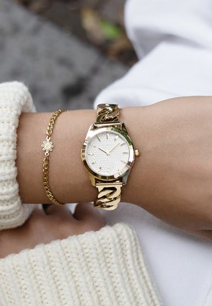 Montre en or avec un cadran texturé blanc, bracelet en maillon doré et délicat bracelet en or avec un petit charme en forme de marguerite, portée sur un poignet habillé d'un pull léger.