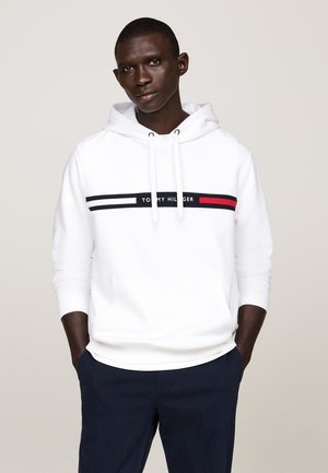 Homme portant un sweat à capuche blanc avec le logo "Tommy Hilfiger" et des rayures bleu marine, associé à un pantalon bleu marine, debout devant un fond uni.