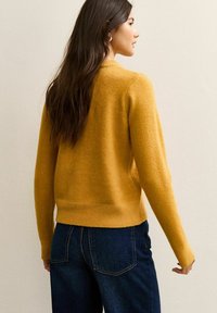 Geel wollen sweater met een ronde hals en ribbelmanchetten, gecombineerd met donkere spijkerbroek. De textuur is zacht en iets pluizig.