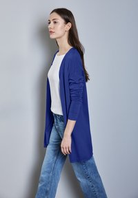 Blå lång cardigan av mjukt tyg, bärs över en vit topp. Matchad med ljusblå utsvängda jeans, poserar modellen mot en neutral bakgrund.