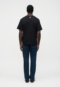 Μαύρο βαμβακερό μπλουζάκι με κοντά μανίκια, κόκκινο σχέδιο στην πλάτη, συνδυασμένο με μπλε τζιν παντελόνι, ευθεία γραμμή, με ορατές λεπτομέρειες ραφών.