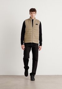 Μπεζ καπιτονέ γιλέκο με φερμουάρ, χωρίς μανίκια, συνδυασμένο με μαύρο πουκάμισο μακρυμάνικο και μαύρο παντελόνι, φορεμένο από μοντέλο σε ουδέτερο φόντο.