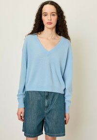 Pull léger bleu avec col en V et manches longues, associé à un short en denim. Texture douce, coupe décontractée et détails de couture visibles.