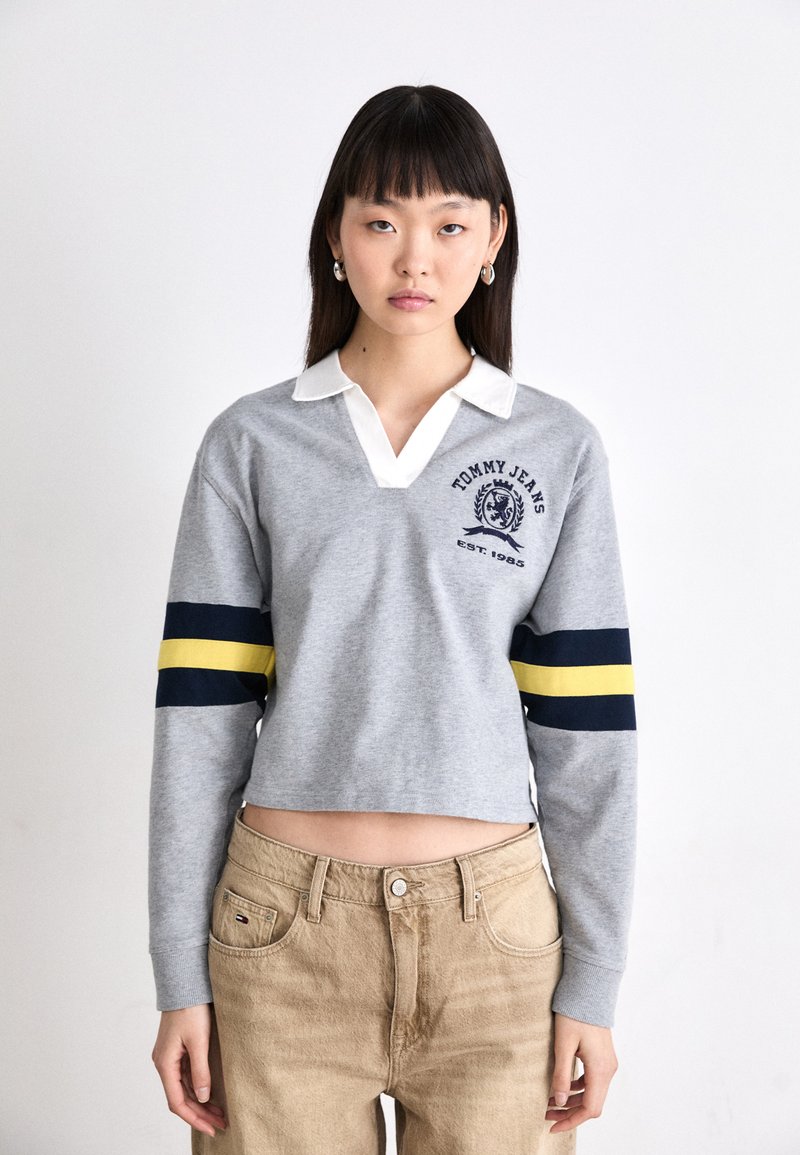 Felpa grigia corta con maniche lunghe, colletto bianco, strisce blu navy e gialle, e logo "TOMMY JEANS" con emblema sul petto.