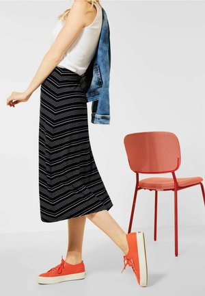 Jupe maxi à rayures noires et blanches, associée à un débardeur blanc et une veste en denim. Le mannequin porte des sneakers orange. Une chaise rouge est visible.