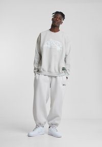 Felpa grigia chiara con stampa grafica, abbinata a pantaloni della tuta coordinati. Sneakers bianche, vestibilità rilassata, polsini a costine e dettaglio con etichetta del logo.