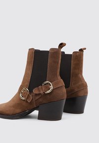 Bottines en suède marron avec des bouts ronds, des panneaux latéraux élastiques et des boucles dorées ; talons carrés épais et tirants à l'arrière.