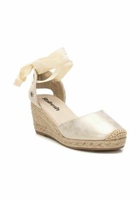 Sandalo con zeppa, superiore in tessuto metallico dorato, soletta in giute intrecciato e punta rotonda. Presenta un cinturino alla caviglia con fiocco per una vestibilità sicura.