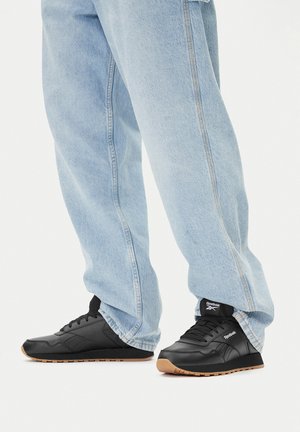 Czarne buty sportowe z teksturowaną skórzaną cholewką i gumową podeszwą w kolorze karmelowym, w połączeniu z jasnoniebieskimi luźnymi dżinsami z widocznym przeszyciem.