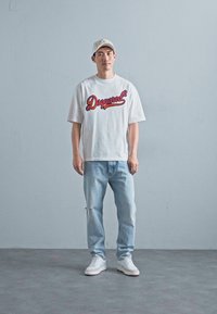 Witte t-shirt met rood "Dsquared" logo, lichtblauwe versleten jeans en witte sneakers, allemaal gedragen met een lichte pet.