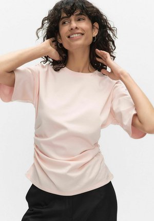 Femme souriante aux cheveux bouclés portant des boucles d'oreilles argentées, un t-shirt rose pâle à manches courtes et un pantalon noir, posant avec les mains près de la tête.