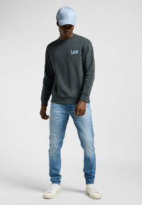 Felpa grigia scura con logo "Lee", cappellino azzurro chiaro, jeans blu sbiaditi e sneakers bianche. Look casual, design minimalista.