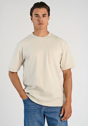 Maglietta beige a maniche corte con scollatura rotonda e tasca sul petto, realizzata in un tessuto morbido, abbinata a jeans in denim blu.