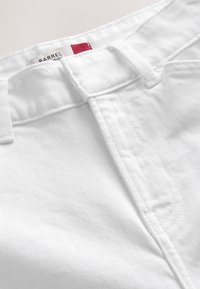 Gros plan sur la taille d'un jean en denim blanc avec passants pour ceinture et étiquette de marque rouge et blanche partiellement visible à l'intérieur.