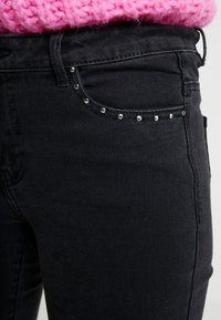 Acercamiento de un bolsillo de jeans de mezclilla negros con pequeñas tachuelas plateadas, usado con un suéter de punto rosa metido por dentro.