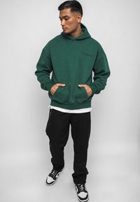 Groene katoenen hoodie met voorzakken, ribbelmanchetten en onderrand. Ggedragen over een wit shirt en gecombineerd met zwarte broek en sneakers.