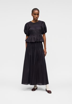 Femme aux cheveux tressés portant un haut noir à manches bouffantes avec volant et une jupe longue assortie, des chaussettes blanches et des ballerines marron foncé.