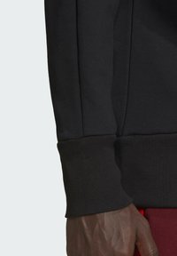 Felpa nera con polsini a coste, caratterizzata da una texture liscia e un design aderente, mostrata su una mano contro uno sfondo a contrasto.