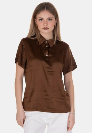 Giovane donna che indossa una polo marrone lucida a maniche corte e pantaloni bianchi, in piedi davanti a uno sfondo chiaro uniforme.