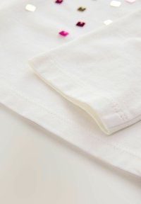 Biała bawełniana koszulka T-shirt złożona, o gładkiej fakturze, z małymi, odblaskowymi, kwadratowymi cekinami w kolorze różowym, złotym i srebrnym, ułożonymi na przodzie.