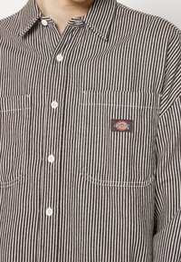 Camicia button-up a righe marroni e bianche con colletto, due tasche frontali e un piccolo logo rettangolare Dickies sulla tasca sinistra.