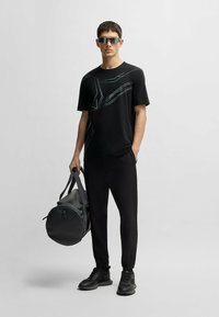 Black t-shirt with white pattern, black pants, and black sneakers. Carrying a round black duffel bag. Silver rectangular sunglasses.